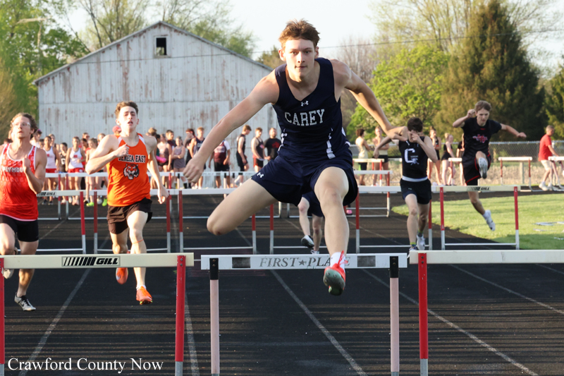 Male hurdler in a navy Carey uniform leaps over a hurdle on a track; other runners and a barn backdrop visible behind him.