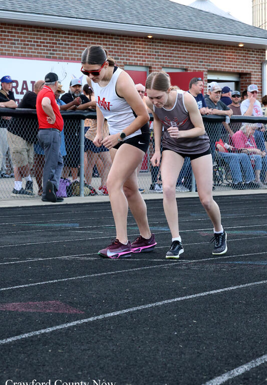 Two female sprinters crouch at the starting line on a track, one in a white top with pink shoes and sunglasses and the other in a gray top; spectators stand behind a chain-link fence.