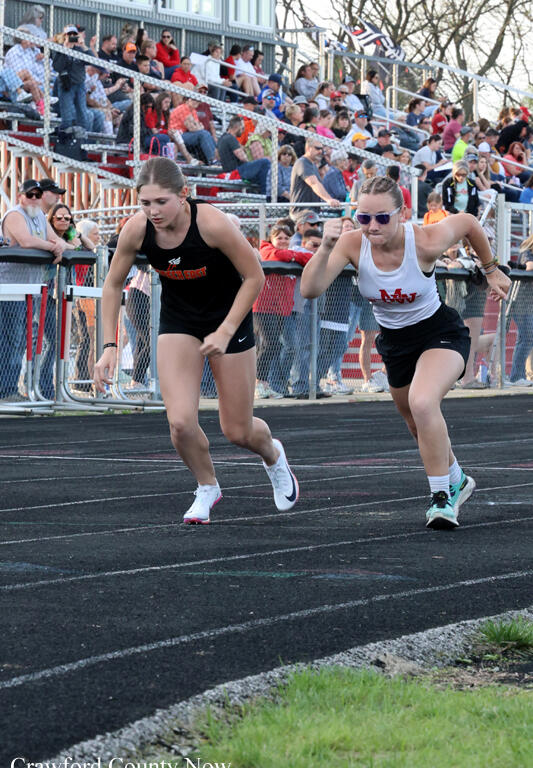 Two young female sprinters sprint from the starting line on a track with a cheering crowd behind a chain-link fence. One wears a black uniform, the other a white top and black shorts.
