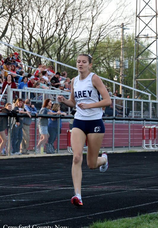 Female high school runner in a white jersey and navy shorts sprinting on a track, with spectators in bleachers behind a chain-link fence.