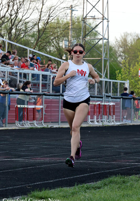 Female runner sprinting on an outdoor track with spectators in the stands behind her.