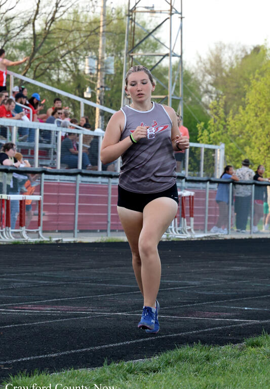 Female sprinter in a gray sleeveless top and black shorts mid-run on a track, with spectators watching from bleachers behind.