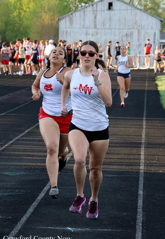 Two female sprinters lead a track race on an outdoor track; the front runner wears a white tank and black shorts with red sunglasses, followed by a teammate in white top and red shorts, with a crowd and barn in the background.