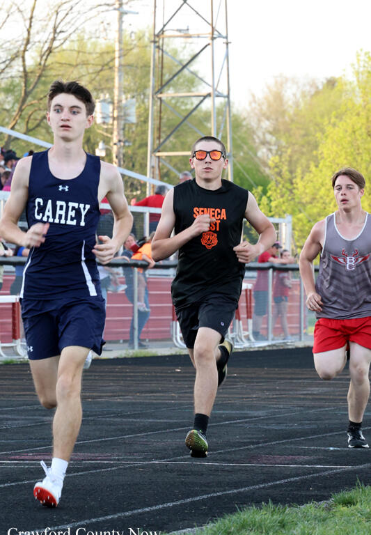 Three high school sprinters race on an outdoor track: left wears navy Carey, center in black Seneca West, right in gray and red. Spectators and bleachers in the background.