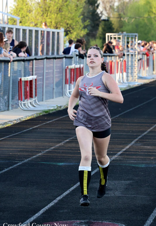 Female sprinter in a gray tank top and black shorts runs on an outdoor track as spectators watch from the sideline fence.