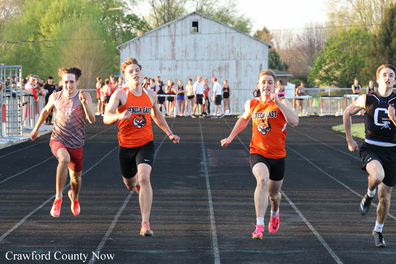 Four male sprinters race down an outdoor track as a crowd watches from the infield, with a weathered barn in the background.