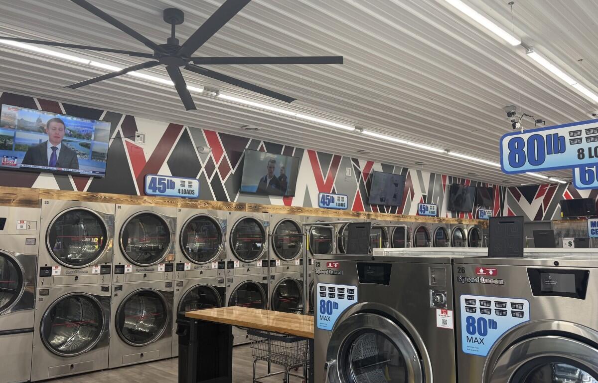 A row of industrial coin-operated front-loading washers lining a laundromat wall, with TVs mounted above and large ceiling fans overhead.