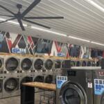 A row of industrial coin-operated front-loading washers lining a laundromat wall, with TVs mounted above and large ceiling fans overhead.