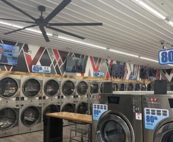 A row of industrial coin-operated front-loading washers lining a laundromat wall, with TVs mounted above and large ceiling fans overhead.