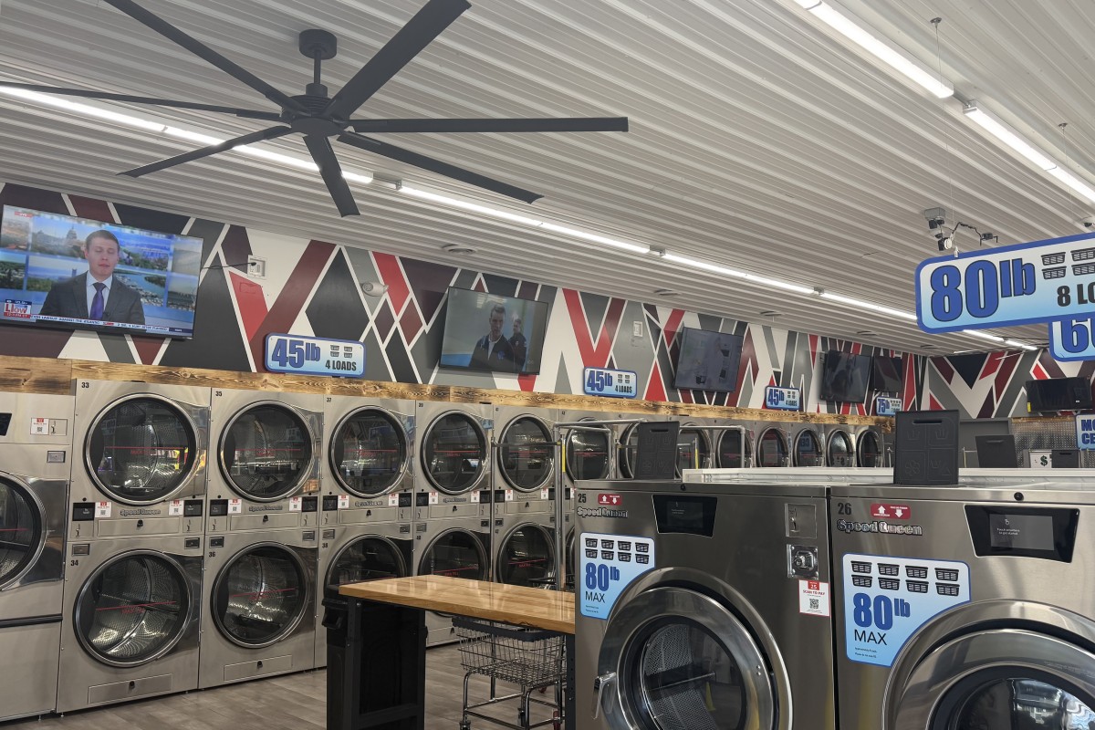 A row of industrial coin-operated front-loading washers lining a laundromat wall, with TVs mounted above and large ceiling fans overhead.