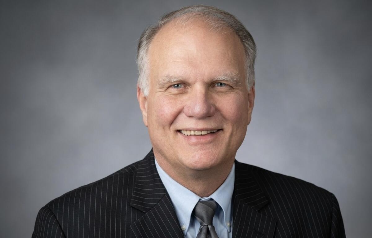 Smiling older man in a dark pinstriped suit, light blue shirt, and gray tie, portrait against a gray gradient backdrop.