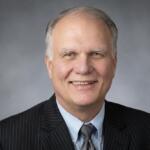 Smiling older man in a dark pinstriped suit, light blue shirt, and gray tie, portrait against a gray gradient backdrop.
