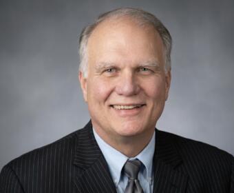 Smiling older man in a dark pinstriped suit, light blue shirt, and gray tie, portrait against a gray gradient backdrop.