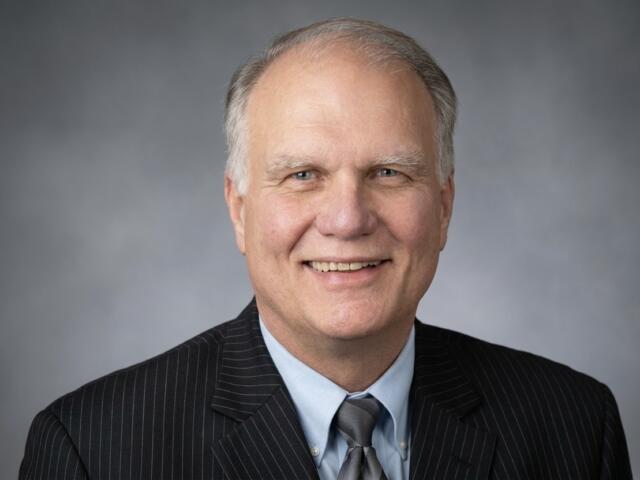 Smiling older man in a dark pinstriped suit, light blue shirt, and gray tie, portrait against a gray gradient backdrop.