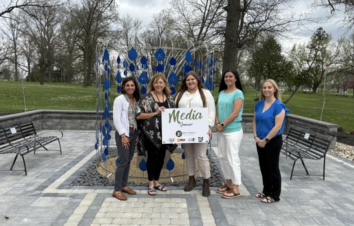 Five women pose together in a park beside a blue teardrop sculpture, holding a 'Media Sponsor' sign.