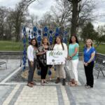 Five women pose together in a park beside a blue teardrop sculpture, holding a 'Media Sponsor' sign.
