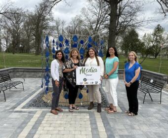 Five women pose together in a park beside a blue teardrop sculpture, holding a 'Media Sponsor' sign.