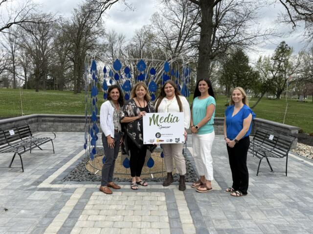 Five women pose together in a park beside a blue teardrop sculpture, holding a 'Media Sponsor' sign.
