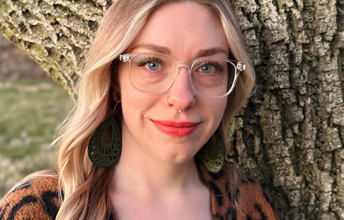 Portrait of a smiling woman with clear glasses, nose ring, green intricate earrings, and a leopard-print top, standing beside a tree trunk outdoors.