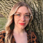 Portrait of a smiling woman with clear glasses, nose ring, green intricate earrings, and a leopard-print top, standing beside a tree trunk outdoors.