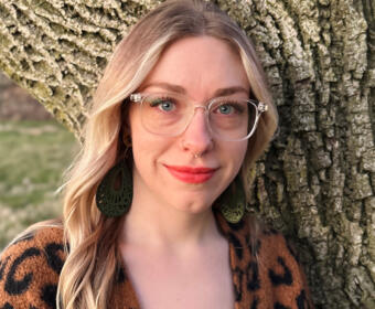 Portrait of a smiling woman with clear glasses, nose ring, green intricate earrings, and a leopard-print top, standing beside a tree trunk outdoors.