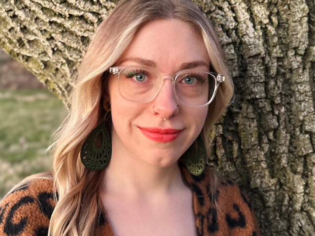 Portrait of a smiling woman with clear glasses, nose ring, green intricate earrings, and a leopard-print top, standing beside a tree trunk outdoors.