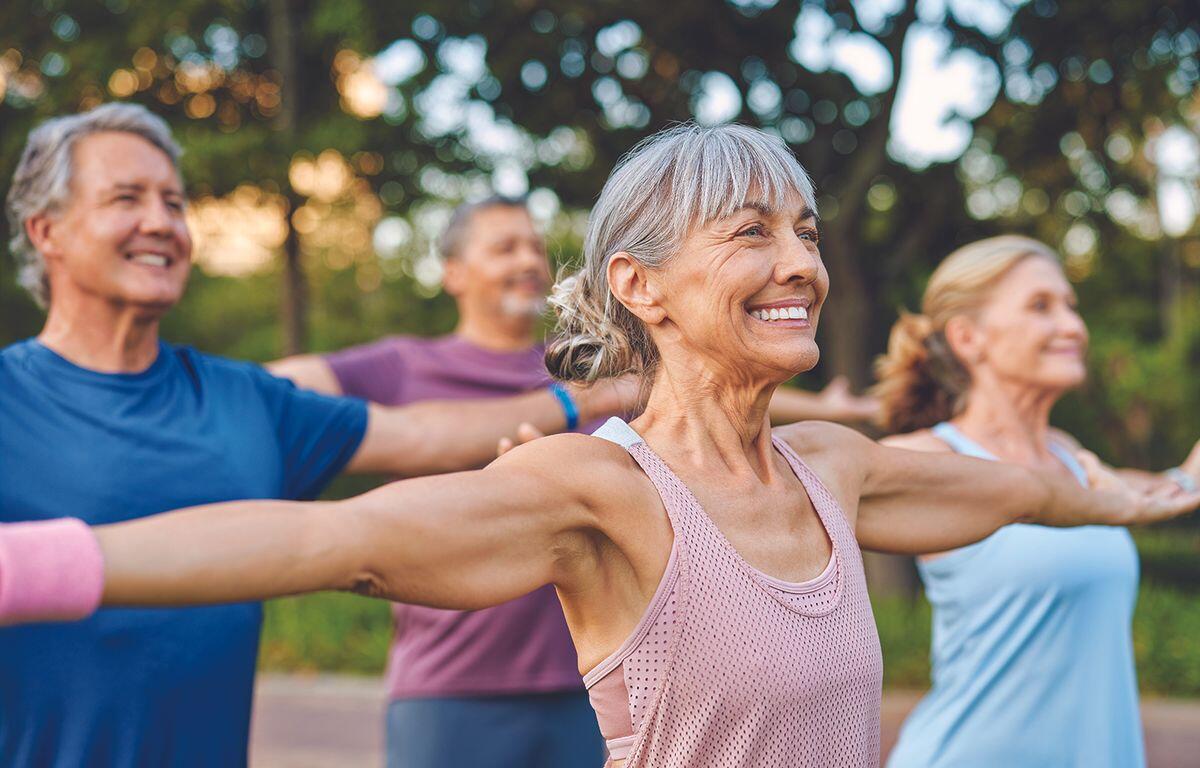 Group of smiling seniors doing arm stretches in a park on a sunny day.