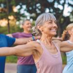 Group of smiling seniors doing arm stretches in a park on a sunny day.