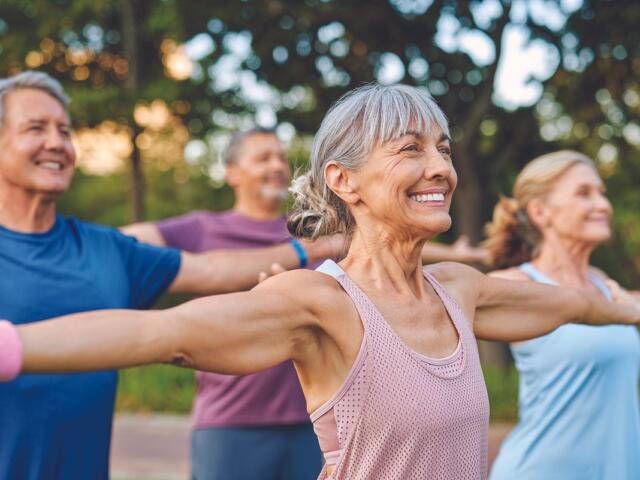 Group of smiling seniors doing arm stretches in a park on a sunny day.