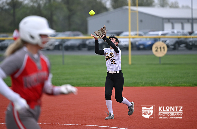 Softball scene: fielder wearing number 22 in a white uniform reaches up to catch a high ball on the infield as a near-blurred runner in red approaches from the left.