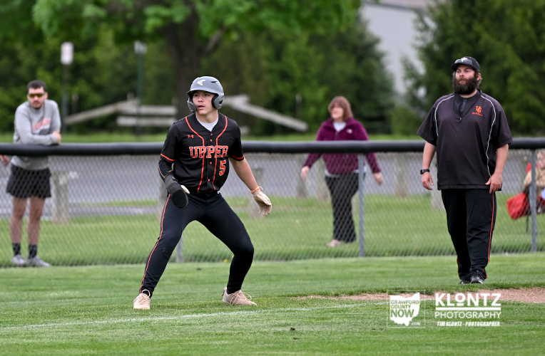 Baseball player in a black and orange uniform with a helmet, standing in the infield and ready to run or bat, with a coach and spectators in the background.