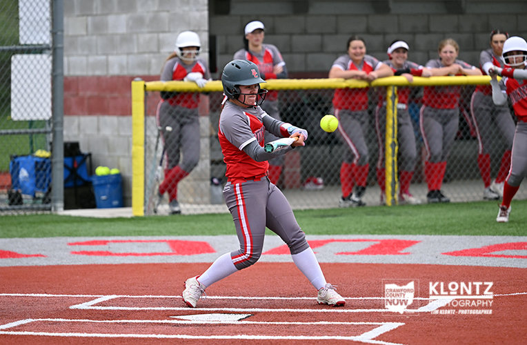 Female softball batter in red and gray swings at a pitched ball during a game on a brown infield, teammates watch from a chain-link fence dugout in the background.