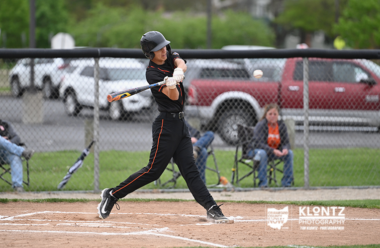 Baseball batter in black uniform swinging at a pitch during a game, with a chain-link fence and spectators in the background.