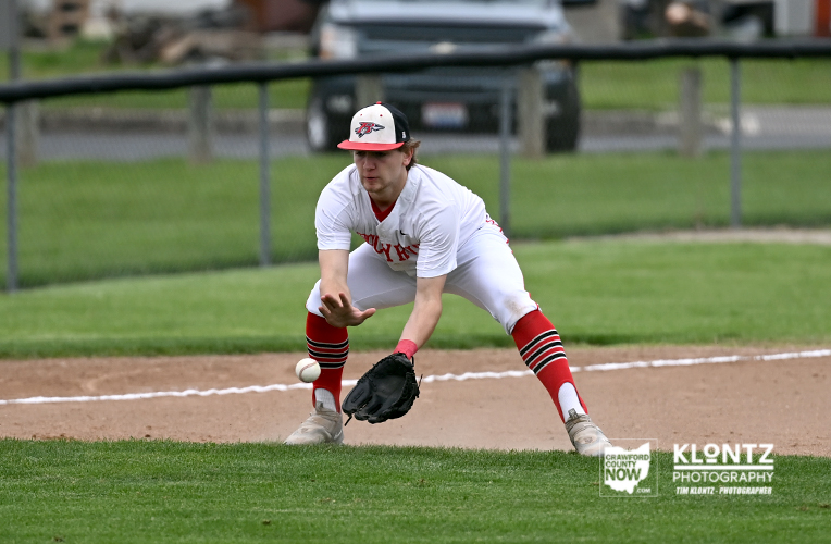 Baseball player in white and red uniform stretches to glove a ground ball on the infield dirt.