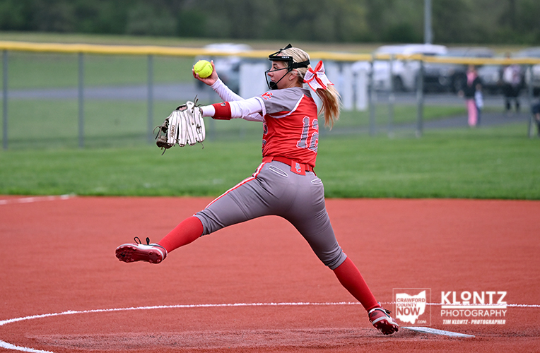 Female softball pitcher in red uniform delivering a pitch on the mound, glove up and ball in hand.