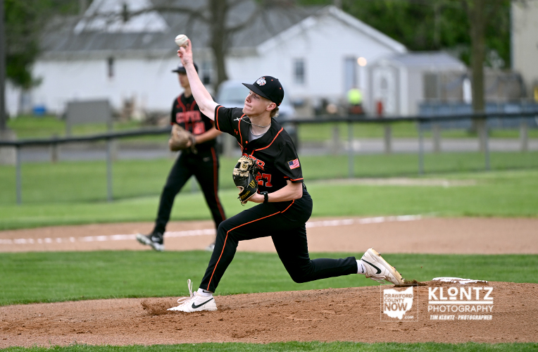 Right-handed baseball pitcher in a black uniform delivering a pitch from the mound with a glove on the left hand and a teammate in the background.