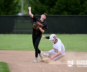 Baseball pitcher in black uniform releases a pitch as a white-uniform runner slides into a base, dust rising on the infield.