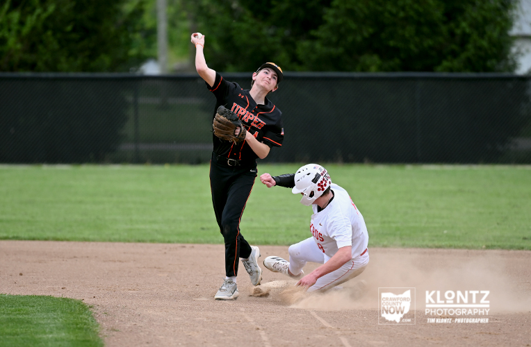 Baseball pitcher in black uniform releases a pitch as a white-uniform runner slides into a base, dust rising on the infield.
