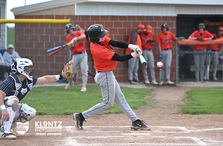 Batter in an orange jersey swings at a pitched ball at home plate, with the catcher in blue gear crouched behind him and teammates watching from the dugout.
