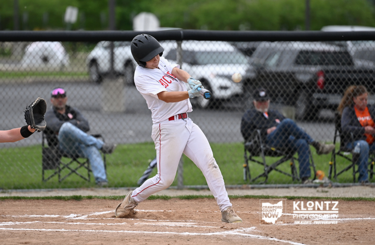 Baseball batter in white uniform swings at a pitch during a game, with a catcher’s glove visible on the left and spectators behind the chain-link fence.