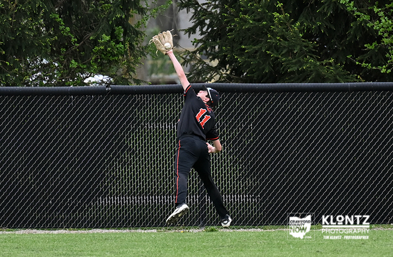 Baseball player in black and red reaching up to catching a ball near a chain-link fence on a grassy field