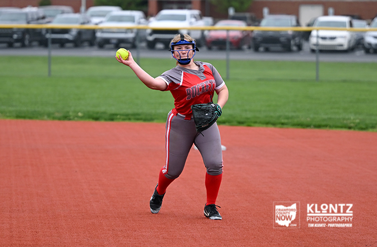 Softball pitcher in a red uniform winds up to throw on a red-dirt field, ball raised in her right hand.