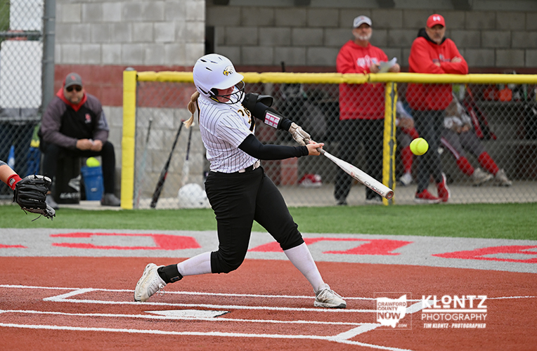 Female softball player in a white pinstriped jersey swings at a pitch on the dirt field, wearing a white helmet and black pants with white socks, as spectators watch behind the fence.