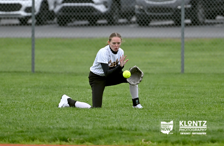 Female softball player in a white and black uniform crouched on the grass, glove up to field a bright yellow ball during a game.
