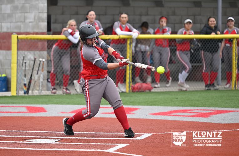 Female softball batter in red and gray uniform swings at a pitch near home plate, helmet on, with dugout and teammates watching behind a chain-link fence.