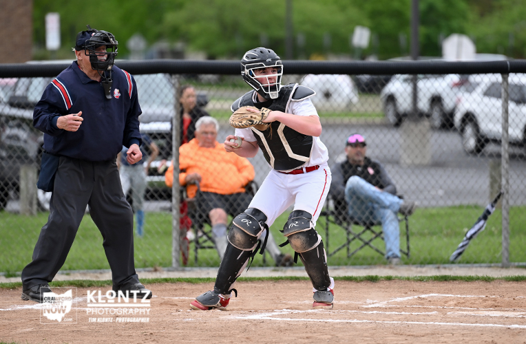 Youth baseball catcher in full protective gear crouches at home plate, awaiting a pitch, with an umpire and spectators behind a chain-link fence.