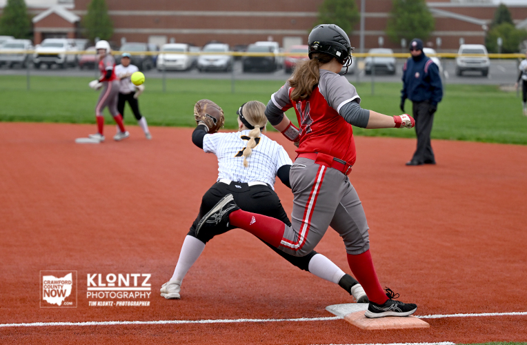 Female softball players in a slide and tag at a base: runner in gray/red slides toward the base as the catcher in white/blue reaches to tag; other players and a fence in the background on an orange infield.