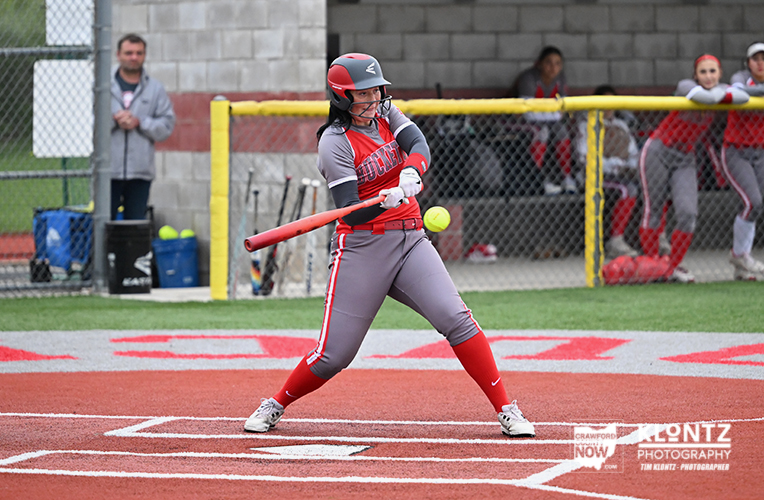 Female softball player in gray and red uniform swinging a bat at a pitch on the field.