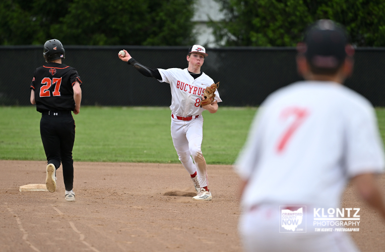 Baseball action on a dirt infield: Bucyrus pitcher in white throws to first as a runner in black (No. 27) moves toward the base; foreground fielder in white No. 7 out of focus.