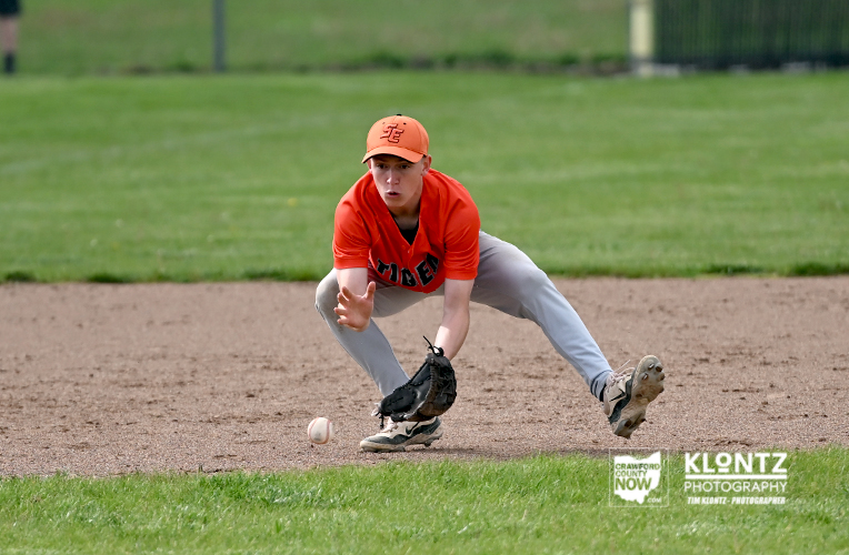 Baseball player in an orange uniform lunges to field a ground ball on a dirt infield, glove reaching toward the ball.